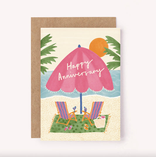 Hand-lettered "Happy Anniversary" sits across a pink striped beach umbrella, surrounded by palm trees, as the sun sets on a pair of cute retro striped beach chairs and some cocktails.