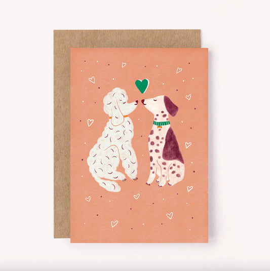 This greeting card features a pair of dogs sharing a kiss. Set on a peachy background with mini hearts surrounding them, this card is perfect for a loved one, Valentines Day or an Anniversary celebration
