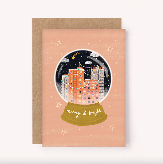 Illustrated "Merry & Bright" holidays card. Set upon a dusty pink background, a festive snow globe is filled with a city skyline and twinkling fairy lights with a sprinkling of snow