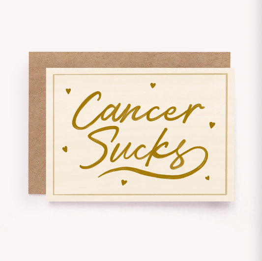 This card was designed to offer support, encouragement, love or just let someone know you're there for them. Illustrated "Cancer Sucks" support card, with gold hand-lettered design on a beige background surrounded by tiny hearts and a simple border