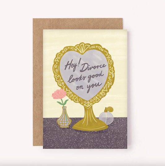 This divorce card features an illustrated ornate gold heart-shaped mirror, which reads "Divorce Looks Good on You". There is a vintage perfume bottle and flower in a vase set upon a purple dressing table